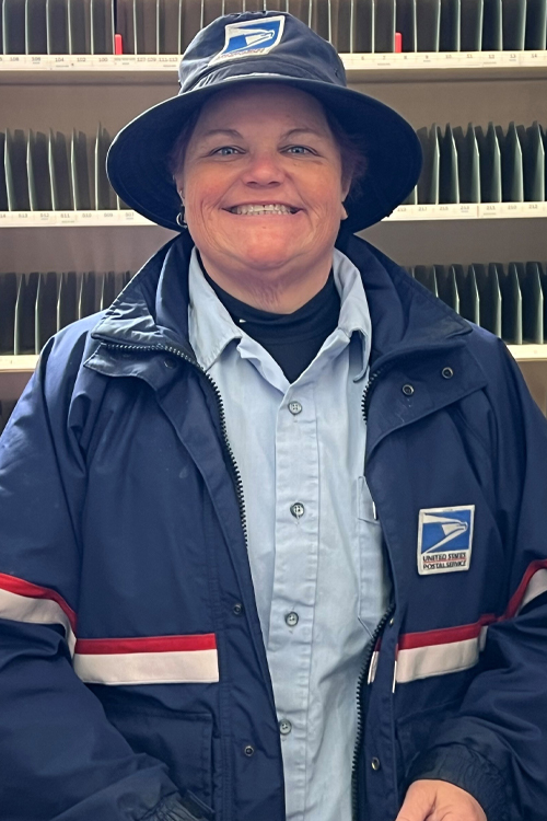 A woman wearing a Postal Service uniform stands and smiles inside a Post Office workroom