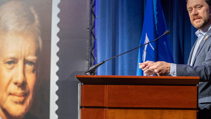 A man in a sport coat stands at a lectern as a poster of the Jimmy Carter stamp looms in the background