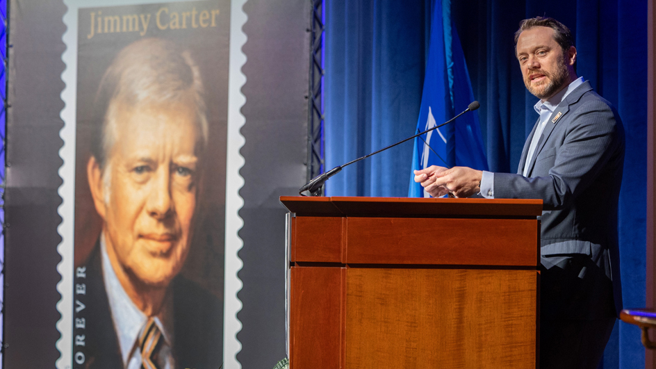A man in a sport coat stands at a lectern as a poster of the Jimmy Carter stamp looms in the background