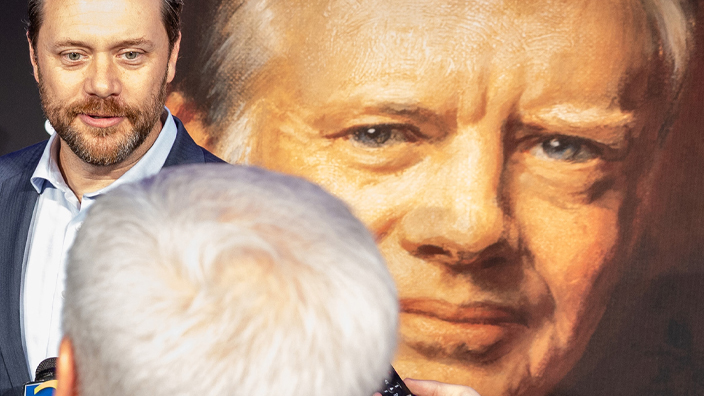 A man in a sport coat stands in front of a poster of the Jimmy Carter stamp and addresses reporters who hold tape recorders