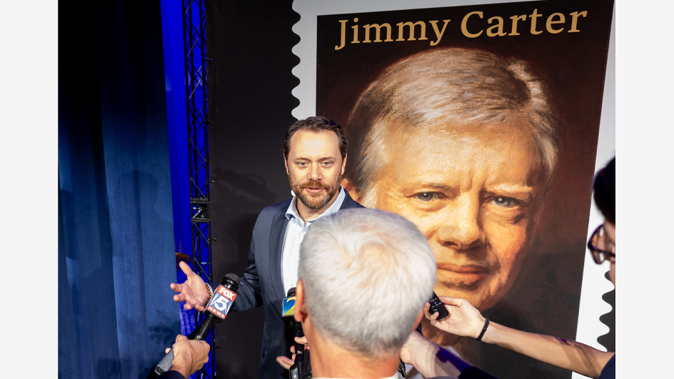 A man in a sport coat stands in front of a poster of the Jimmy Carter stamp and addresses reporters who hold tape recorders