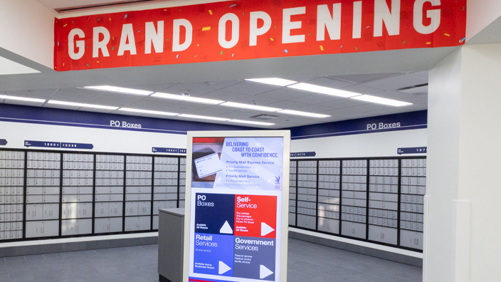 A clean, modern Post Office lobby with a “grand opening” sign hanging above a bank of PO Boxes