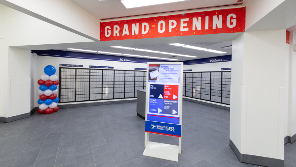 A clean, modern Post Office lobby with a “grand opening” sign hanging above a bank of PO Boxes