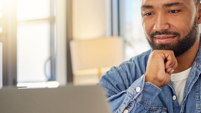 A man sits at a dining room table and looks at a laptop