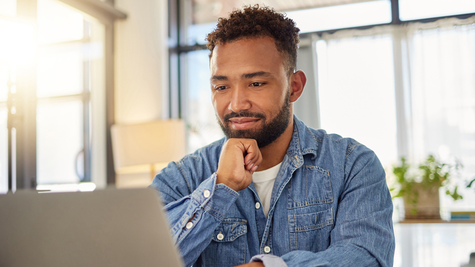 A man sits at a dining room table and looks at a laptop