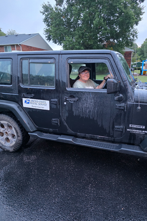 A woman sits in a Jeep with a Postal Service decal on a rear passenger door