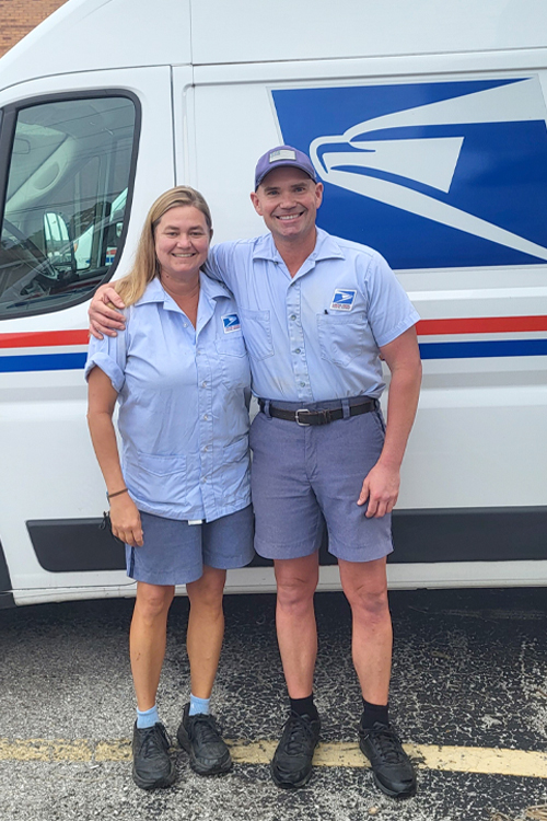 A woman and a man wearing postal uniforms stand next to a USPS delivery vehicle