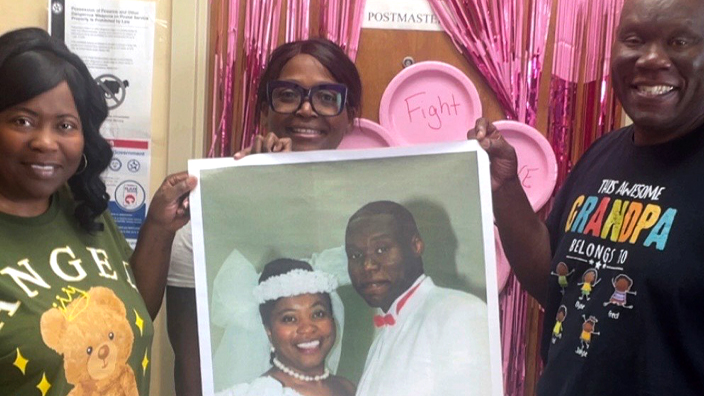 Three people stand together in a Post Office retail lobby; a woman in the middle holds an enlarged wedding photo of a smiling couple
