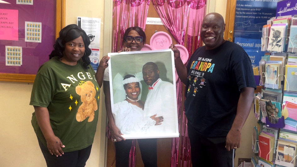 Three people stand together in a Post Office retail lobby; a woman in the middle holds an enlarged wedding photo of a smiling couple