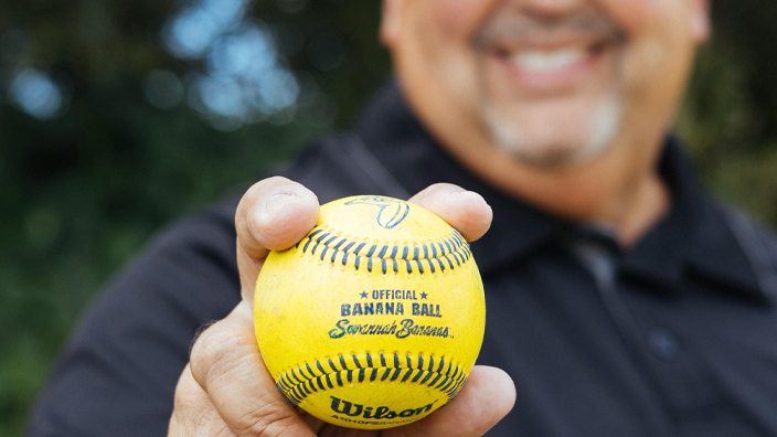 A smiling man stands in a field and holds a bright yellow baseball bearing the words “Savannah Bananas”