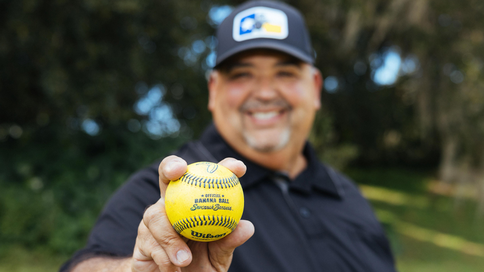 A smiling man stands in a field and holds a bright yellow baseball bearing the words “Savannah Bananas”