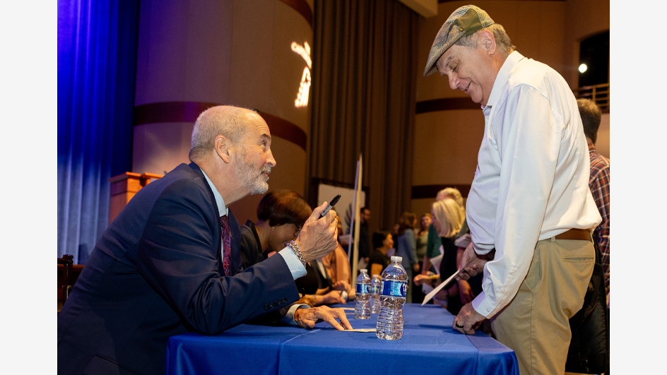 A smiling man in a business suit sits at a table and signs a document for a man who stands near him