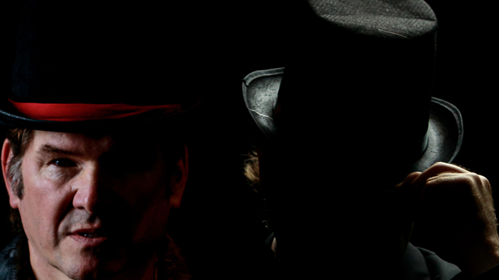 A man dressed in an 1880s costume stands in a darkness, shadow partially obscuring his face