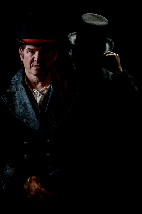 A man dressed in an 1880s costume stands in a darkness, shadow partially obscuring his face