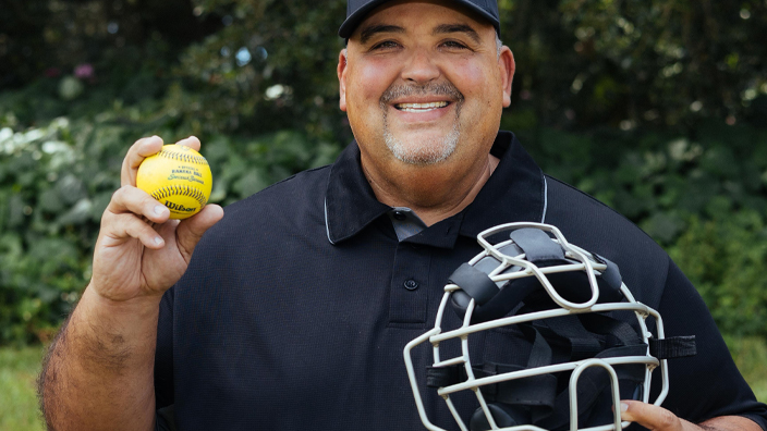 A smiling man stands in a field and holds an umpire’s mask and a yellow baseball