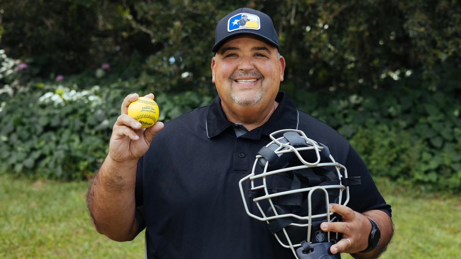 A smiling man stands in a field and holds an umpire’s mask and a yellow baseball