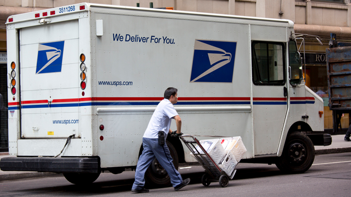 A postal worker pushes a load of packages on a hand truck away from a USPS delivery vehicle.