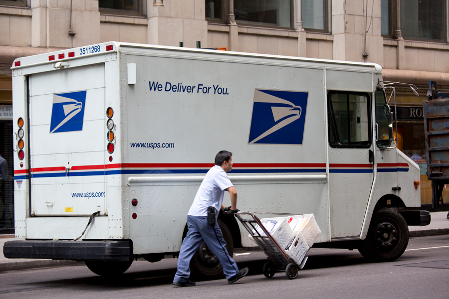 A postal worker pushes a load of packages on a hand truck away from a USPS delivery vehicle.