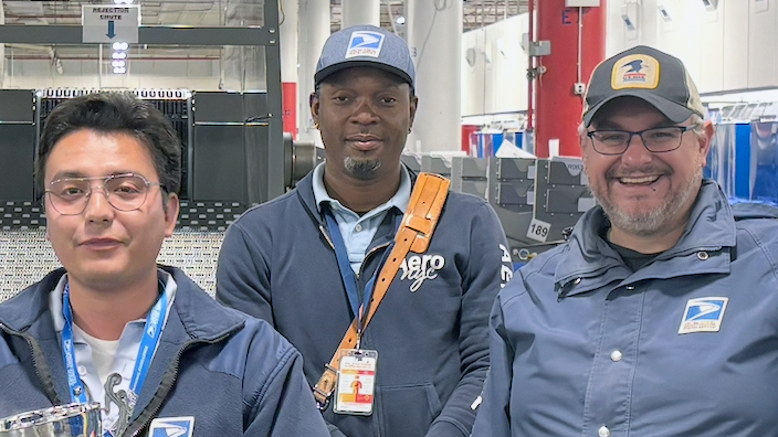 Five people gather around a trophy inside a postal processing plant