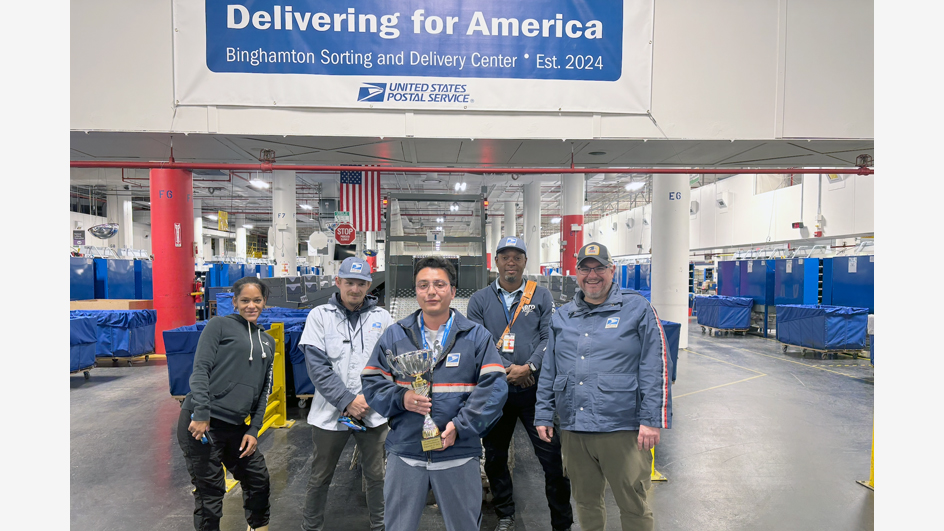Five people gather around a trophy inside a postal processing plant