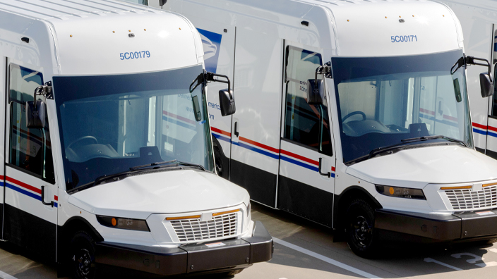 A row of new-style USPS delivery trucks are parked on a lot