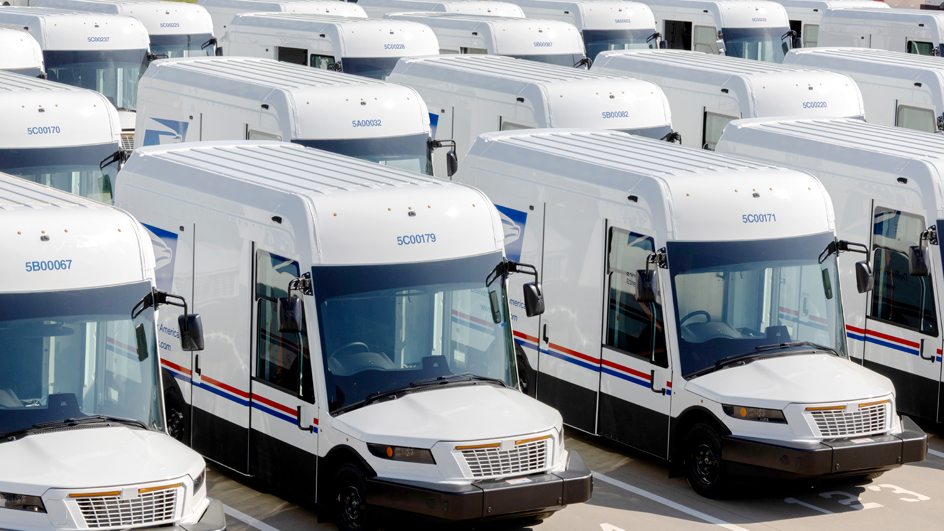 A row of new-style USPS delivery trucks are parked on a lot