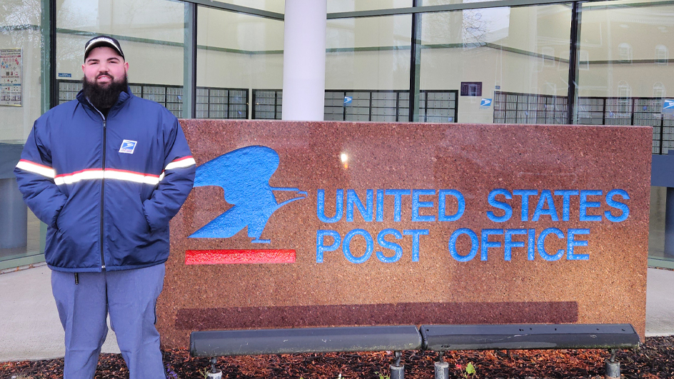 A bearded man wearing a USPS jacket stands next to a U.S Post Office sign