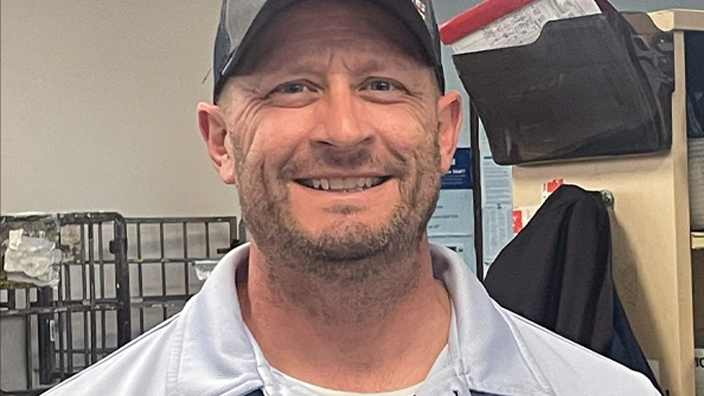 A smiling man wearing a Postal Service uniform stands in a Post Office workroom