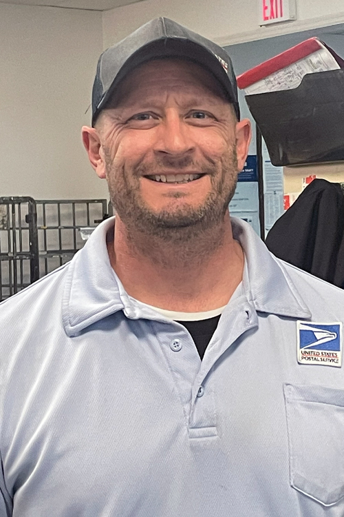 A smiling man wearing a Postal Service uniform stands in a Post Office workroom