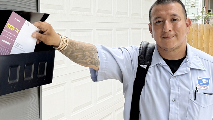 A young man wearing a Postal Service uniform stands near a mailbox on the front porch of a house