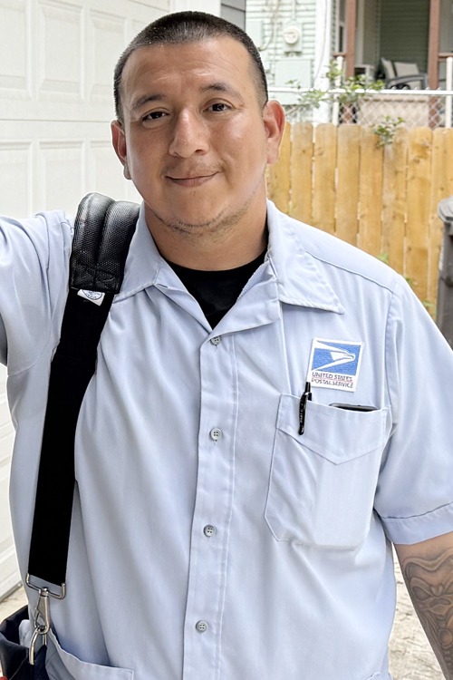 A young man wearing a Postal Service uniform stands near a mailbox on the front porch of a house