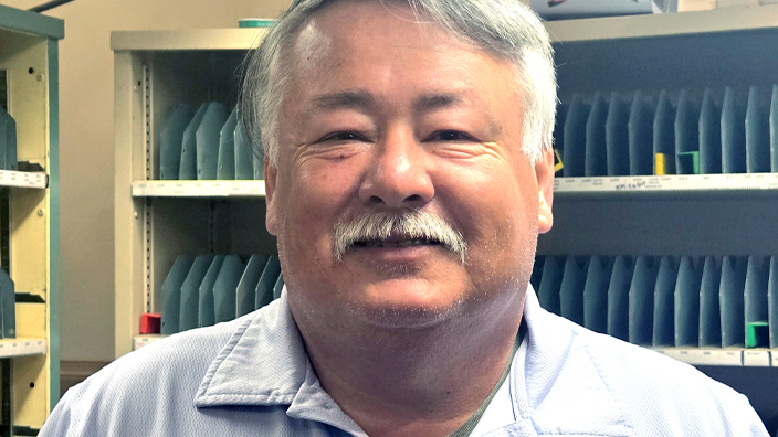 A man wearing a Postal Service uniform stands in a Post Office workroom