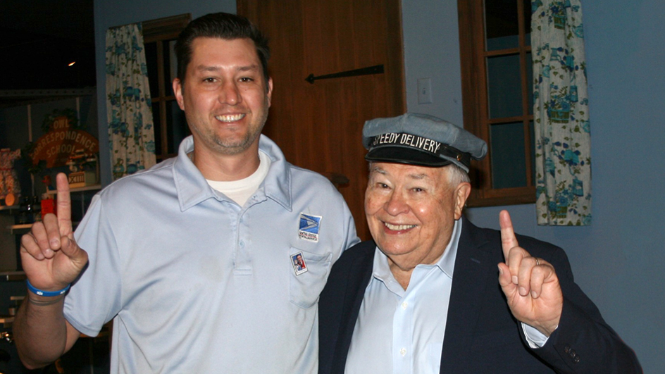 A man in a postal uniform stands next to an older man