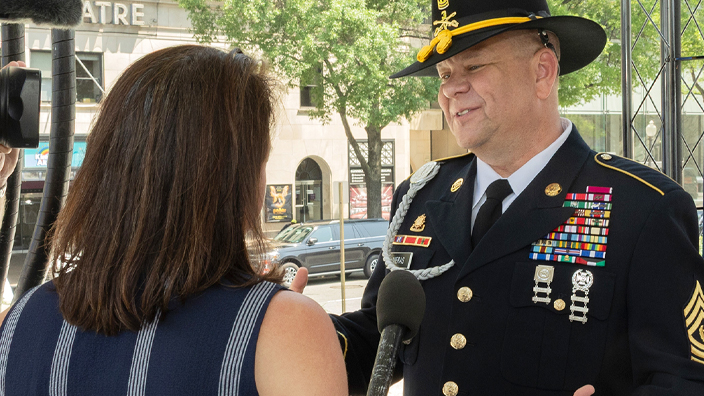 A man in a military uniform stands next to a poster of military-themed stamps and speaks to a video crew