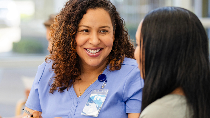 A female doctor in scrubs sits and speaks to a patient in a hospital waiting room