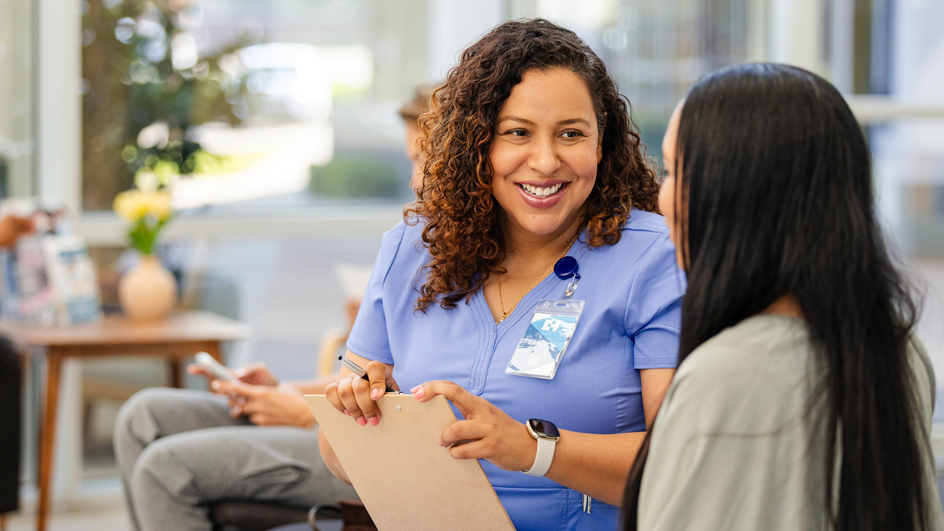 A female doctor in scrubs sits and speaks to a patient in a hospital waiting room