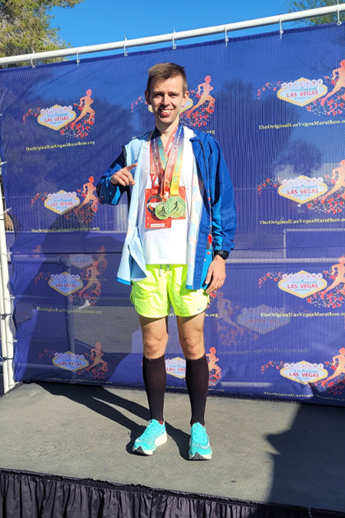 A man in running garb stands on a platform and smiles with two medals around his neck