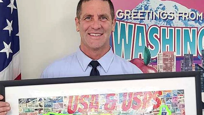 A smiling man wearing a postal uniform holds a framed map of the United States made of stamps