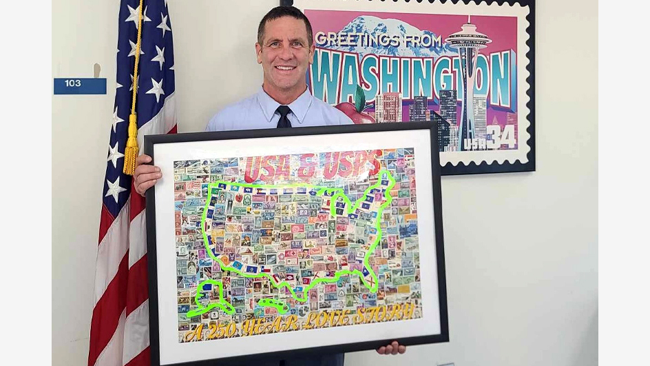 A smiling man wearing a postal uniform holds a framed map of the United States made of stamps