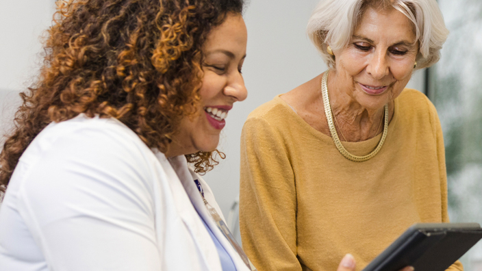 A smiling doctor stands in a medical examining room and shows a tablet screen to a patient sitting on a table