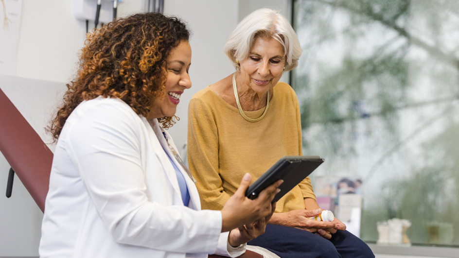 A smiling doctor stands in a medical examining room and shows a tablet screen to a patient sitting on a table