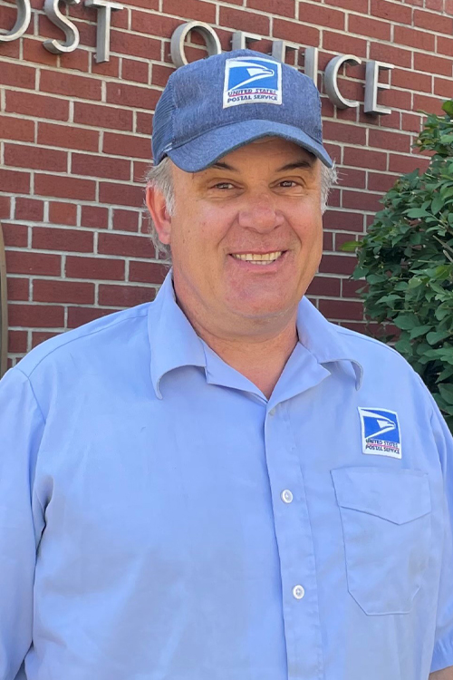 A man in a postal uniform stands outside a brick Post Office building