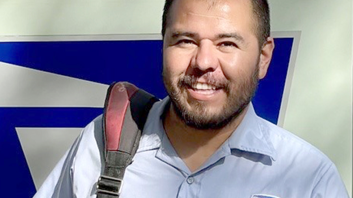 A man wearing a postal uniform stands by a postal delivery vehicle