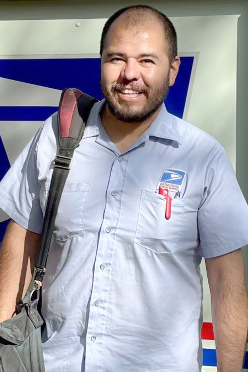 A man wearing a postal uniform stands by a postal delivery vehicle