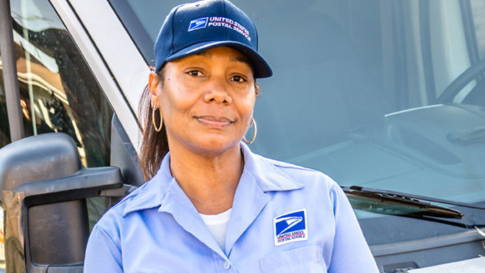 A woman wearing a Postal Service uniform smiles and leans against a delivery vehicle