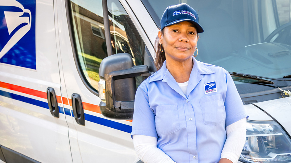 A woman wearing a Postal Service uniform smiles and leans against a delivery vehicle