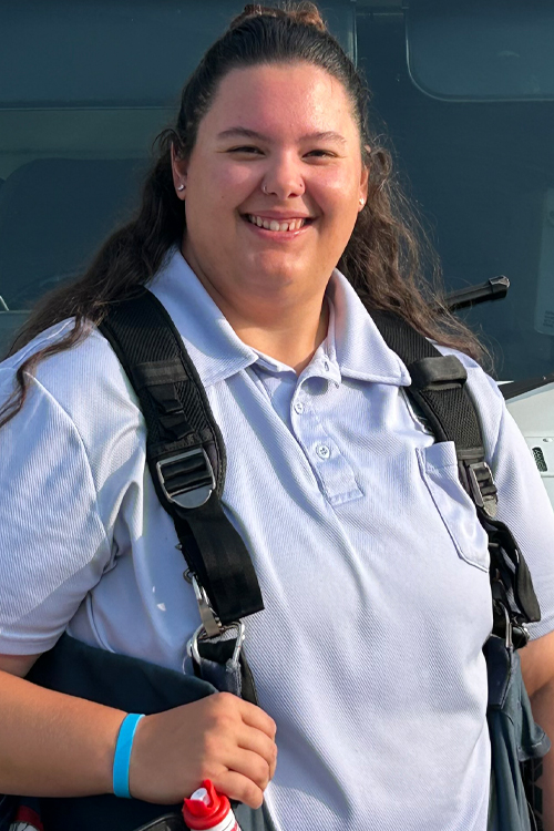 A woman in a postal uniform smiles while standing near a USPS delivery vehicle