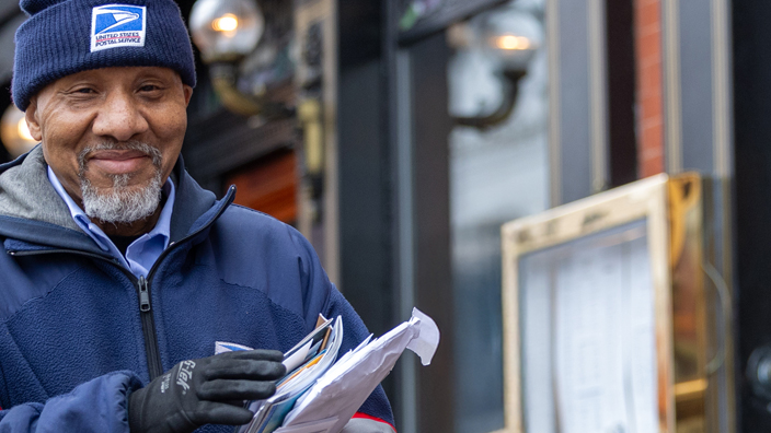 A smiling letter carrier walks along a sideway near a retail shop flanked by two large, colorful toy soldier decorations