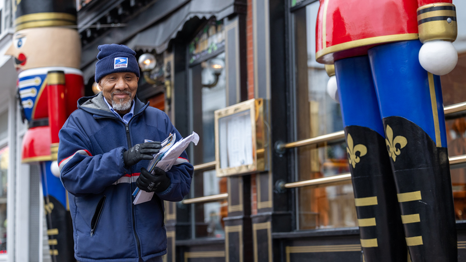 A smiling letter carrier walks along a sideway near a retail shop flanked by two large, colorful toy soldier decorations