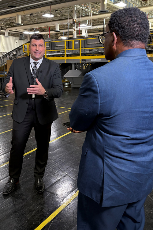 A man in a suit stands on the workroom floor of a USPS processing plant and addresses a TV reporter